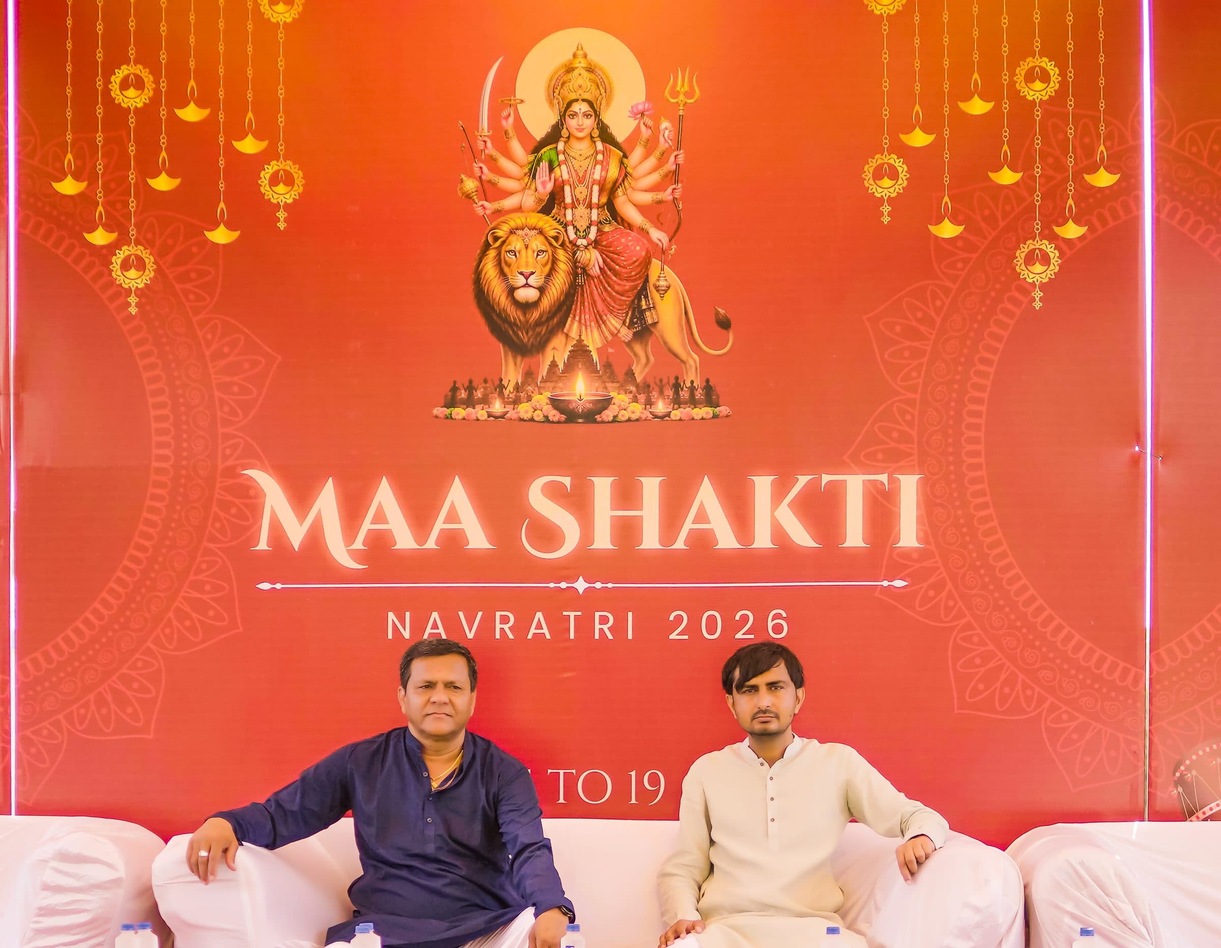 The two organizers of Maa Shakti Navratri, seated in front of the festival backdrop showing Maa Ambe.