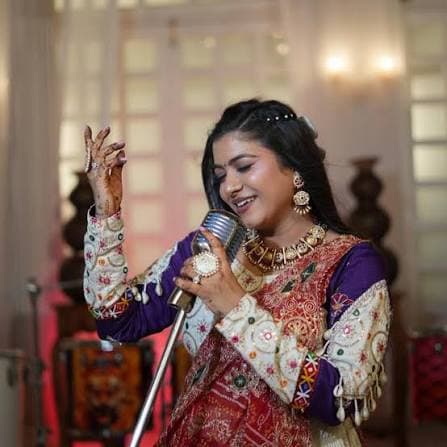 Jaya Patel in a purple sleeved outfit with a red embroidered dupatta and gold jewellery, singing into a microphone in a warmly lit room.