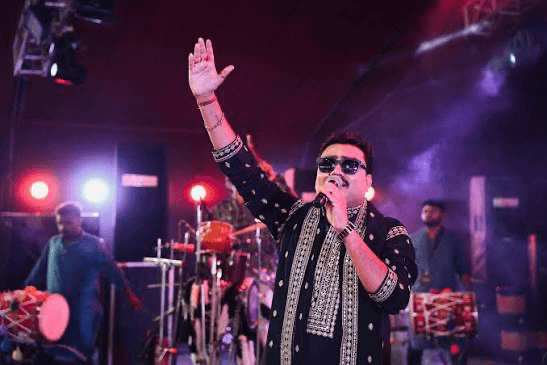 Jignesh Barot in a black embroidered jacket and sunglasses, singing into a microphone with one hand raised, with dhol players behind him under red and blue stage light.