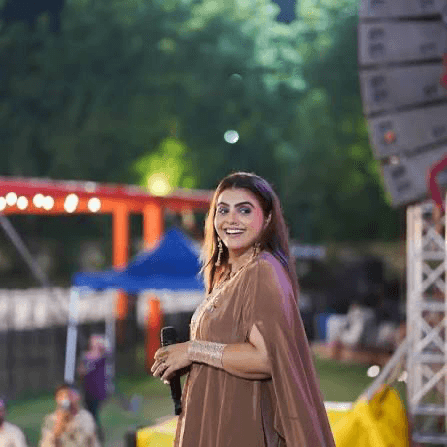Kajal Maheriya in a copper coloured draped outfit and long earrings, smiling and holding a microphone at an open air venue with festival lights behind her.