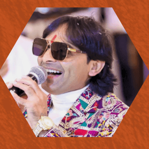 Kamlesh Barot in sunglasses and a brightly patterned kediyu, singing into a microphone.