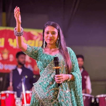 Neha Suthar in a green embroidered outfit with one arm raised and a microphone in the other hand, in front of drums on stage.