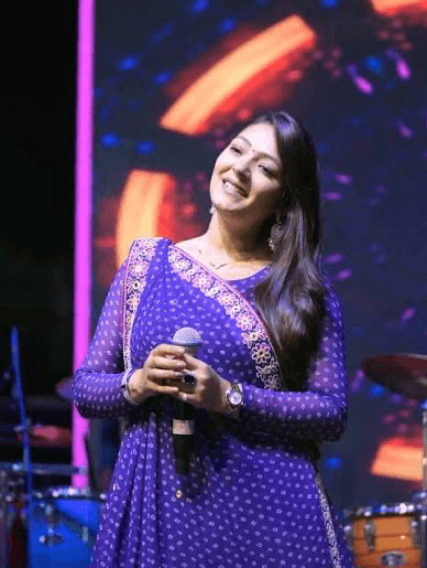 Priti Patel in a purple dotted saree, smiling with her head tilted and a microphone in her hands, in front of a bright stage screen.