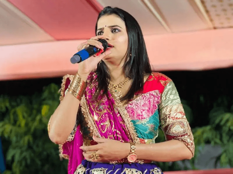 Rajal Barot in a pink, purple and gold embroidered outfit, singing into a blue microphone.