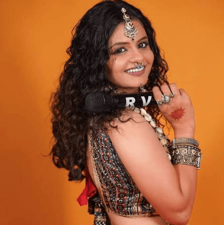 Riddhi Vyas with dark curly hair and a gold headpiece, smiling against an orange backdrop with a microphone marked RV on her shoulder.