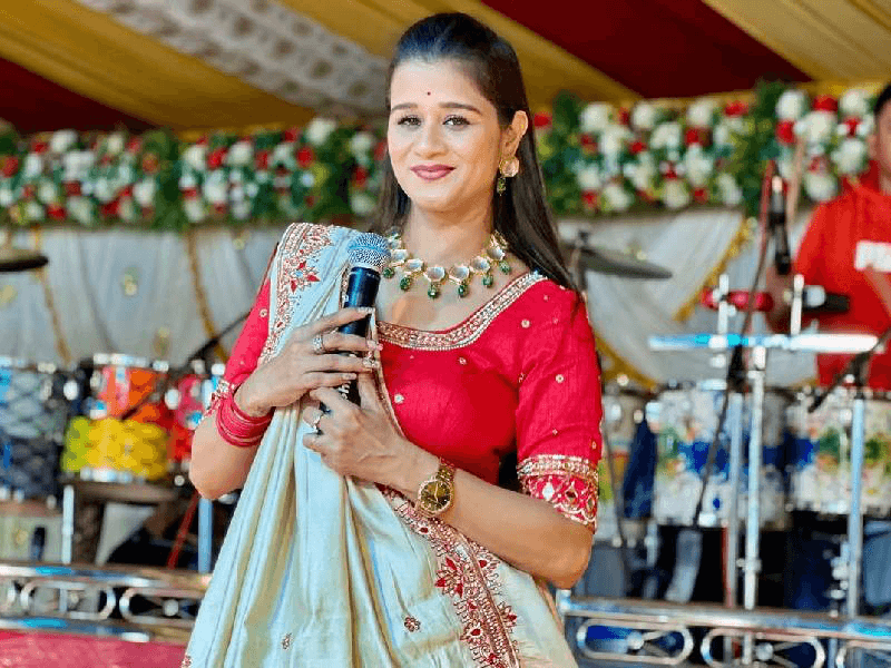 Trusha Rami in a red embroidered blouse and cream saree with a gold necklace, holding a microphone in front of a flower decorated stage and dhols.